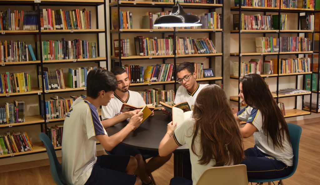 Alunos sentados em mesa redonda dentro da biblioteca prontos para treinar inglês