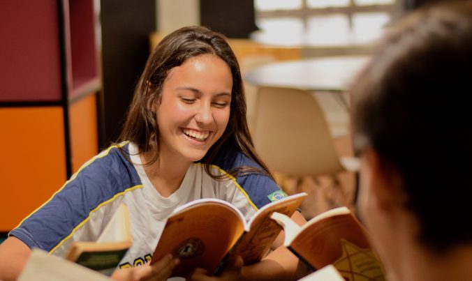 Jovem sorrindo e lendo um livro dentro da biblioteca mostra que o hábito de ler é também prazeroso