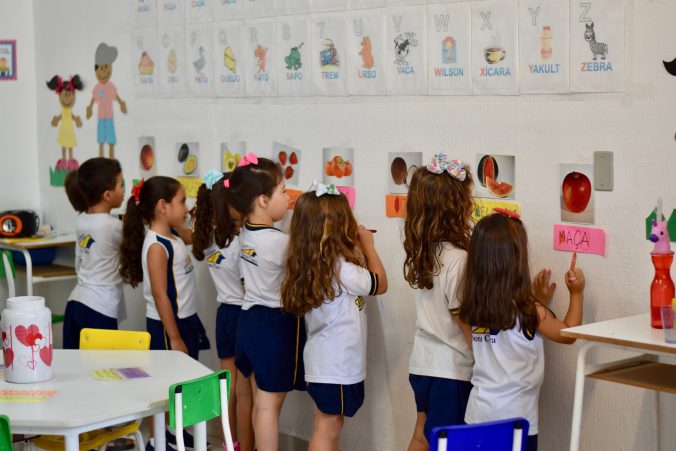 Crianças na sala de aula, em pé, de frente para a parede, escrevendo o nome das frutas abaixo das gravuras correspondentes
